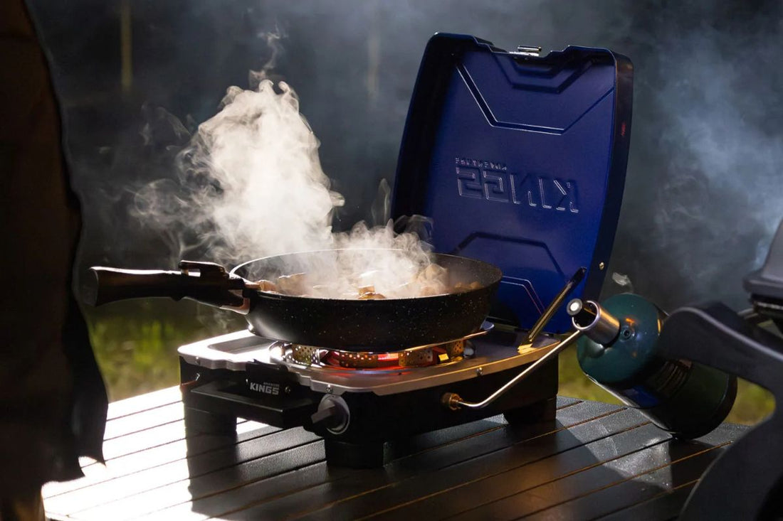 Portable camping stove cooking food in a pan with steam rising, set up on an outdoor table beside a gas canister