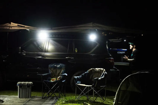 Night campsite with vehicle-mounted lights, camping chairs, and outdoor gear for overlanding